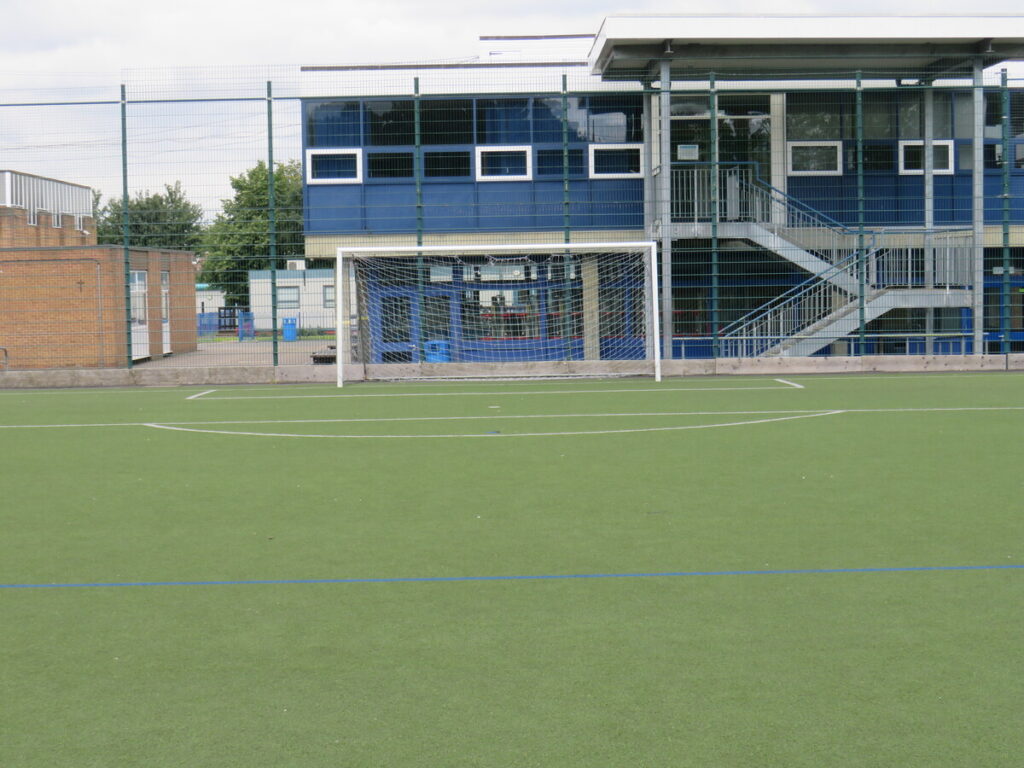 School fenced football astro turf pitch facing goal. With school buildings in background.