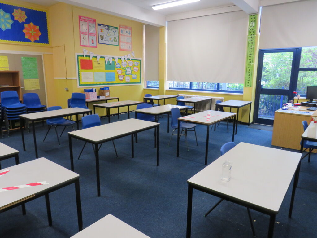 Class room with tables laid out for single pupil/student use. Red and white striped tape crossed on desk tables. Walls decorated with hand craft paper and useful information notes, posters and forms. Teachers desk, glass door fire exit and windows in shot.