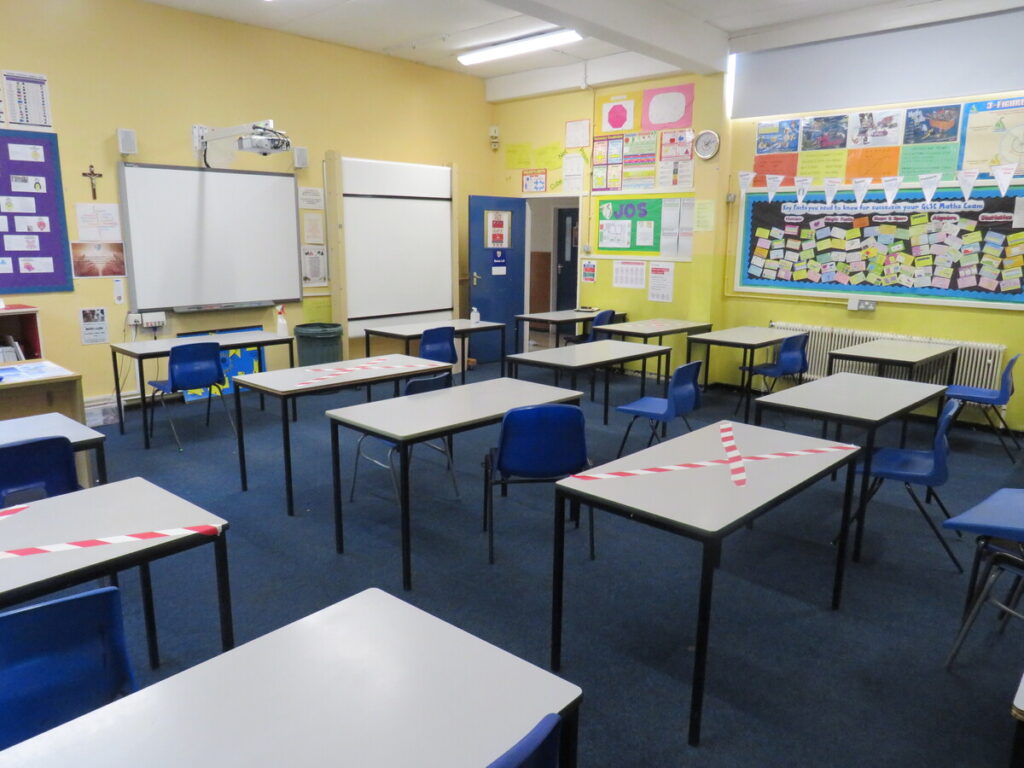 Class room with tables laid out for single pupil/student use, facing front class room at whiteboard and teachers desk. Red and white striped tape crossed on desk tables. Walls decorated with hand craft paper and useful information notes, posters and forms.