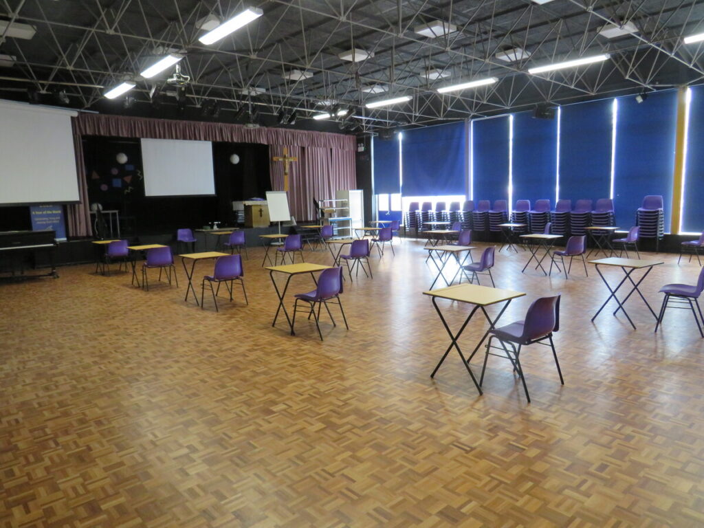 School hall with tables and chairs set out in rows ready for use, facing school stage with a cross displayed, whiteboard, piano and projector screens. School benches and chairs stacked and organised to side by windows.