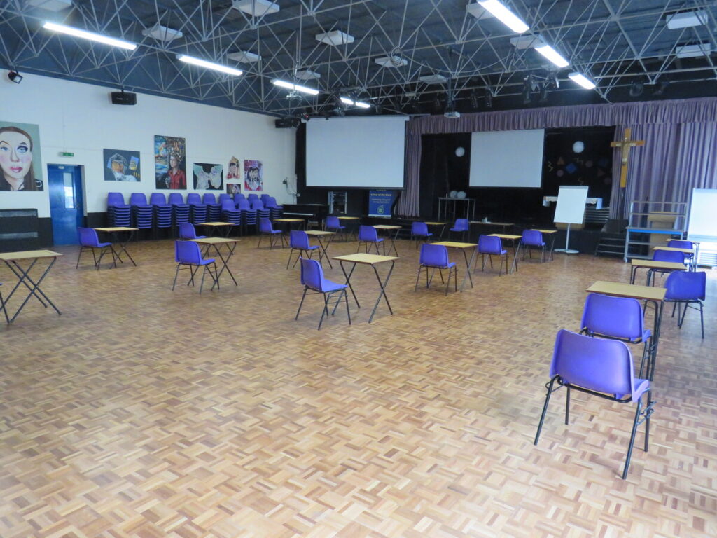 School hall with tables and chairs set out in rows ready for use, facing school stage with a cross displayed, whiteboard and projector screens. Display of art paintings on the school hall walls. school benches and chairs stacked and organised to side by hall walls and windows.