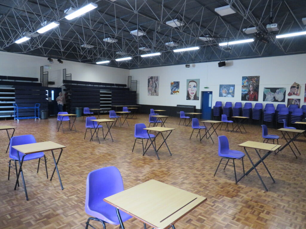 School hall with tables and chairs set out in rows ready for use. Display of art paintings on the school hall walls. school benches and chairs stacked and organised to side by hall walls.