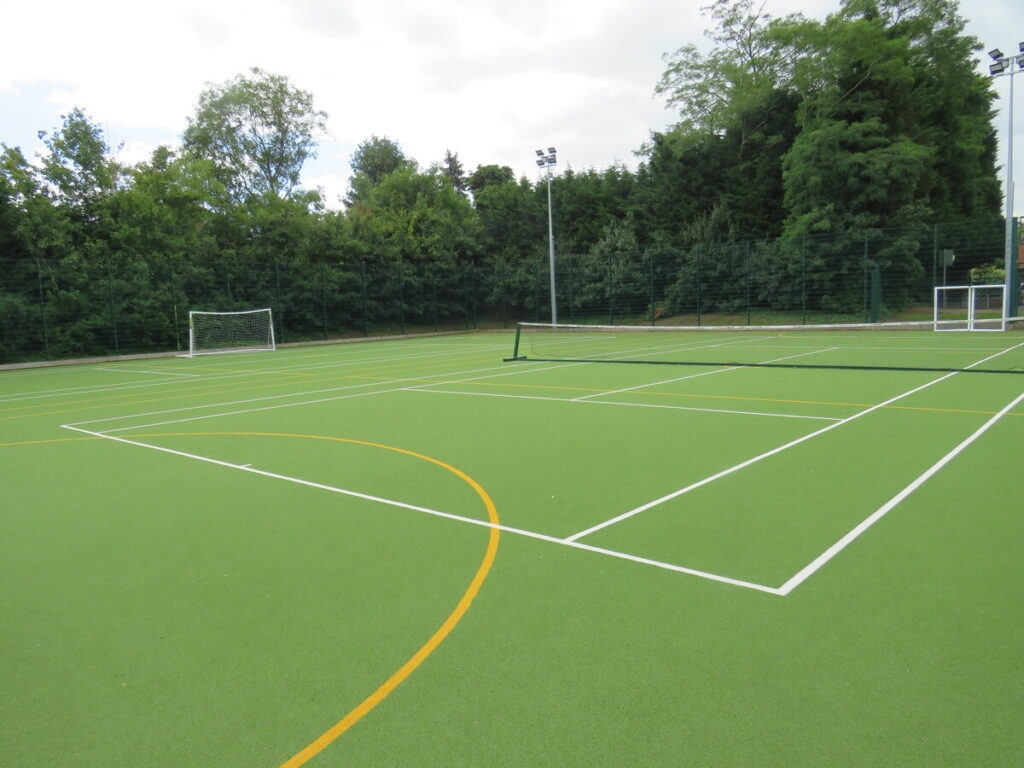 School's outdoor astro turf football pitch, tennis court, with flood light and trees in background.