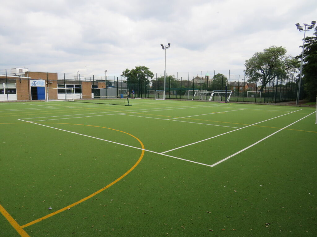 School's outdoor astro turf football pitch, tennis court and netball court, with flood light. School building, trees and houses in background.