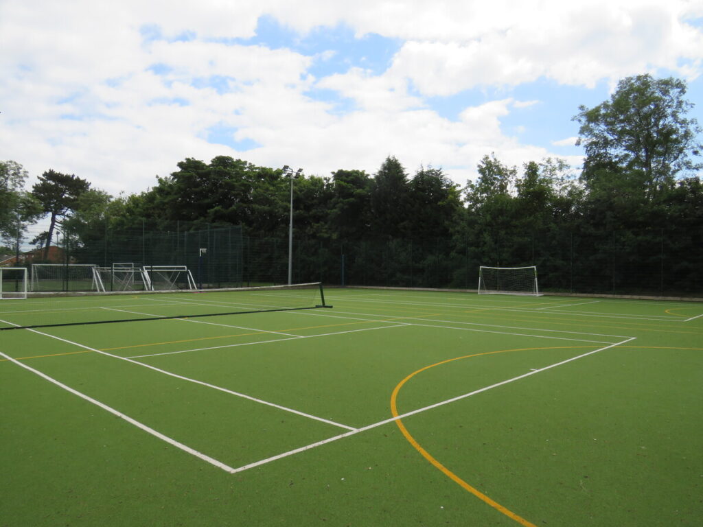 School's outdoor astro turf football pitch, tennis court and netball court, surrounded by trees.