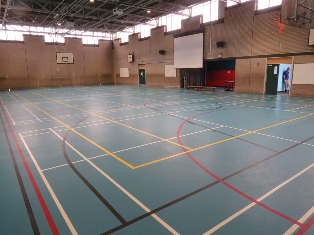 Archbishop Ilsley Catholic school and sixth form school's sports hall, with basketball hoops, projector screen, football goals and playing courts/pitch. Staff member in door way.