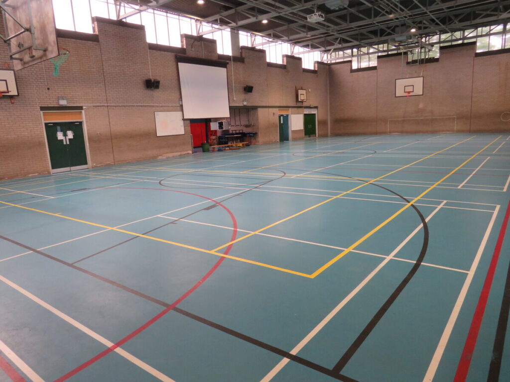 Archbishop Ilsley Catholic school and sixth form school's sports hall, with basketball hoops, projector screen, football goals and playing courts/pitch.