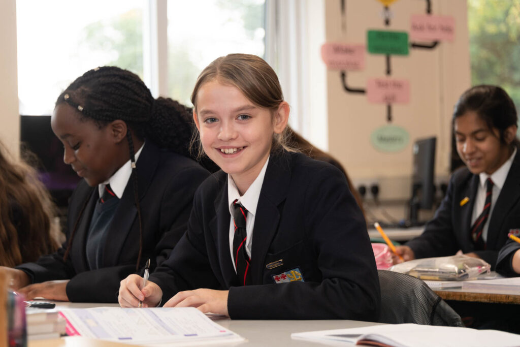 Class of pupils sitting down at desk's in classroom with open books looking forward to front of classroom, writing down notes.