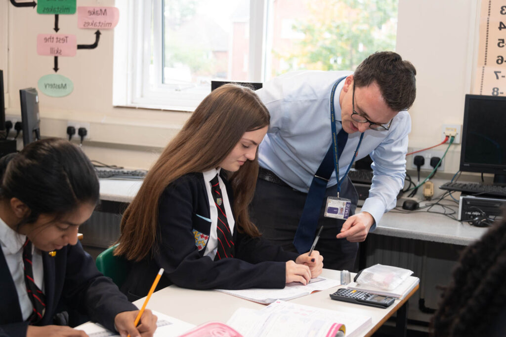 Class of pupils sitting down at desk's in classroom working, with assistance from teacher.