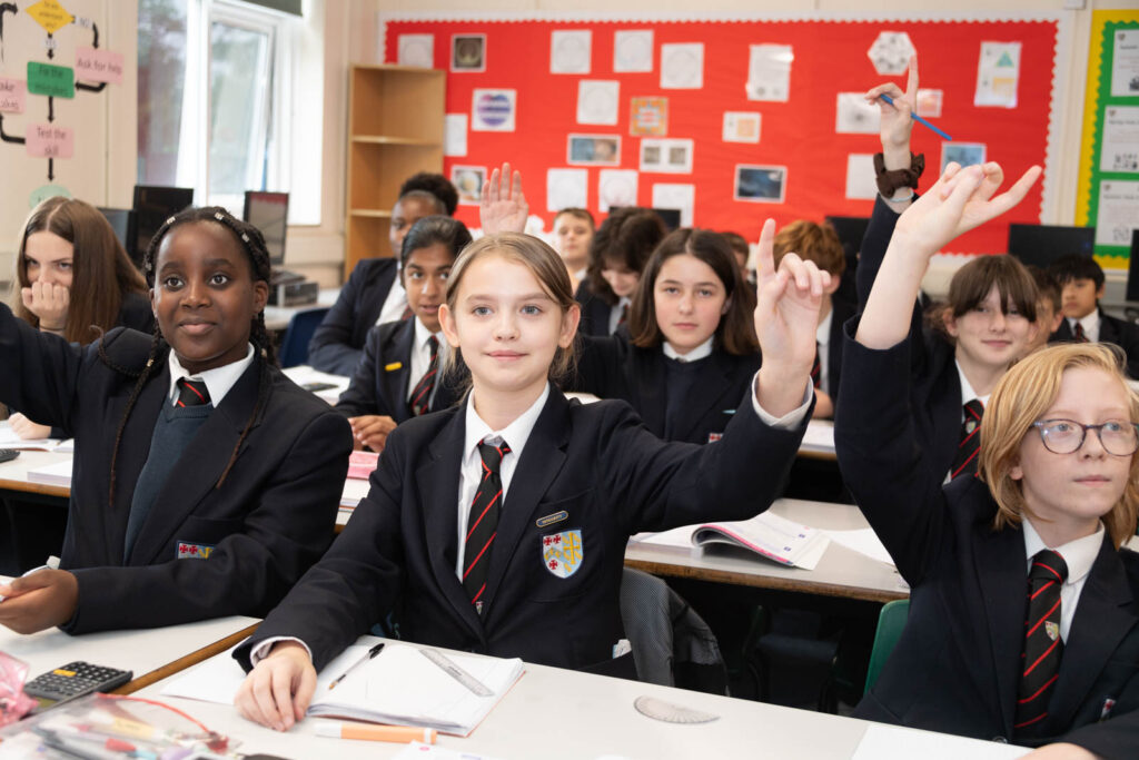 Class of pupils sitting down at desk's in classroom with hands up, facing the forwards.