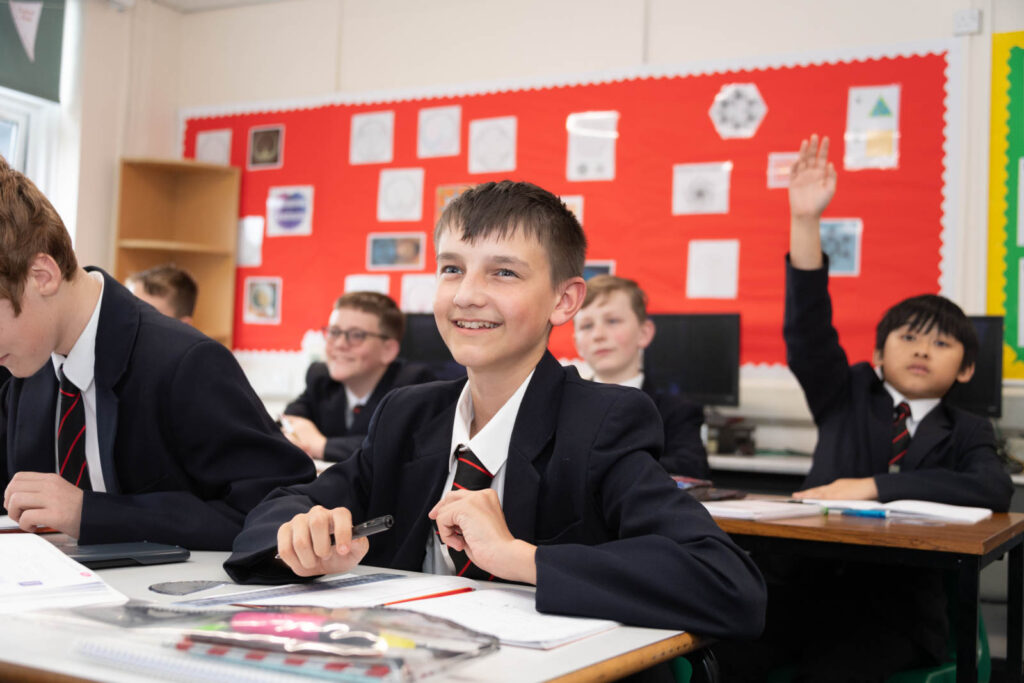 Pupils sitting down at desk's in classroom with open books writing notes, looking towards front of classroom. pupil in background looking forward with hand in the air to ask question.