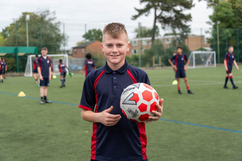 Pupils/students outside on football pitch training. Pupil holding football facing forward posture, other pupils training in the background.