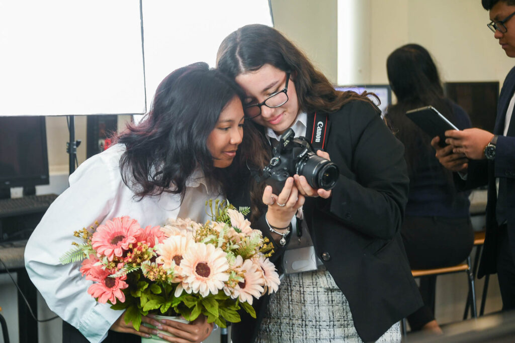 Pupil/student holding a camera showing the other student a photo. While other students work in background. Studio lights in bacground.