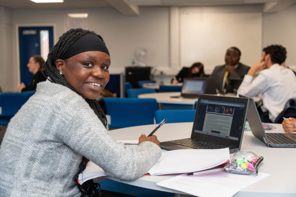 Six form pupils/students working at desks/tables, writing down work in books and laptop.