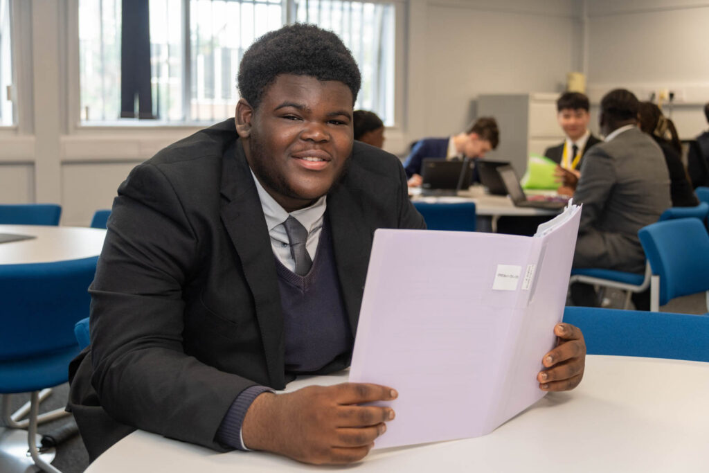 Six form pupil reading out work folder on table/desk. Background pupils/students working around desks/tables, writing down work and on laptops, in work space/class room.