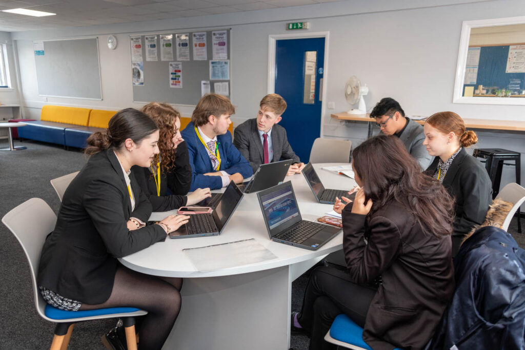 Six form pupils/students working at around desk/table, writing down work and on laptops, in work space/class room.