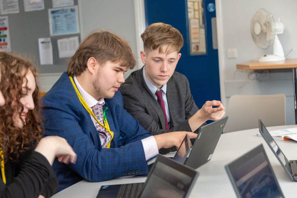 Six form pupils/students working at desk/table, writing down work and on laptops, in work space/class room.