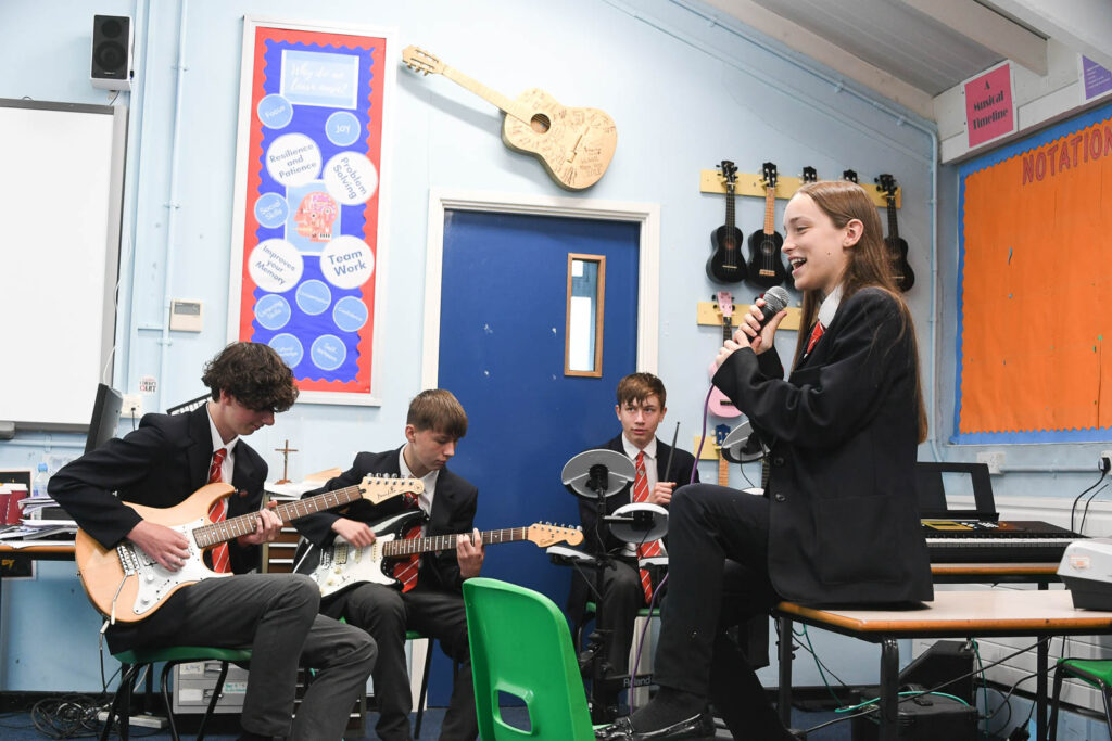 Music class room pupils having band practice jam. Pupil holding microphone singing, sitting on table. The pupils in background playing on guitar and drums.