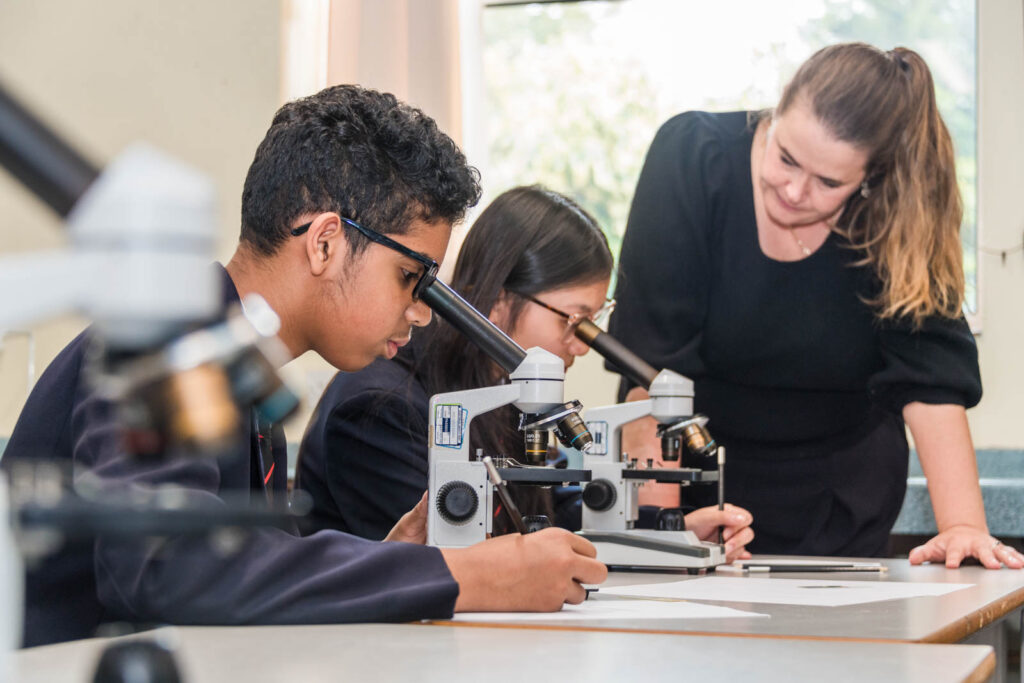 Pupils in science class at desk/table writing down information, while looking down microscope. With teacher watching over them.