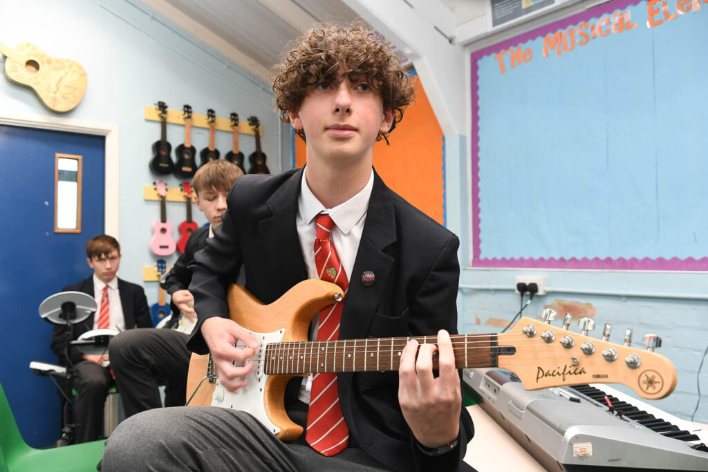 Music class room pupils having band practice jam. Pupils playing on guitar. The pupil in background playing on drums.