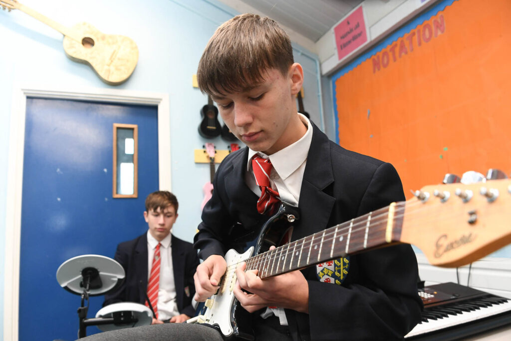 Music class room pupils having band practice jam. Pupil playing guitar. The pupil in background playing on drums.