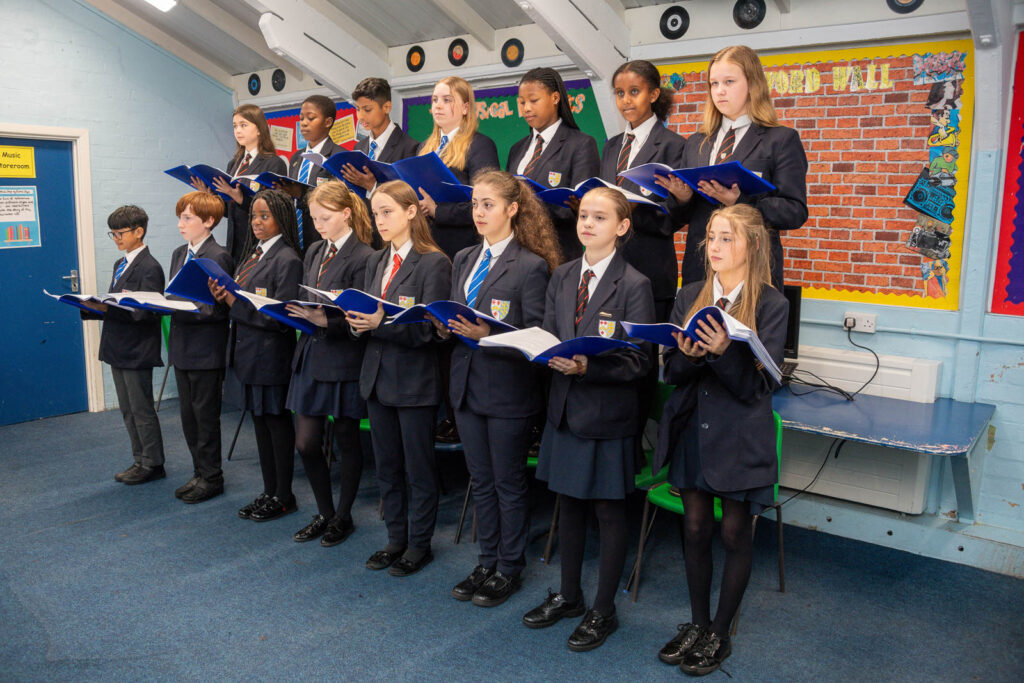 Whole class pupils singing in class, with lyric books in hand. Pupils in two rows, back pupils standing on chairs.