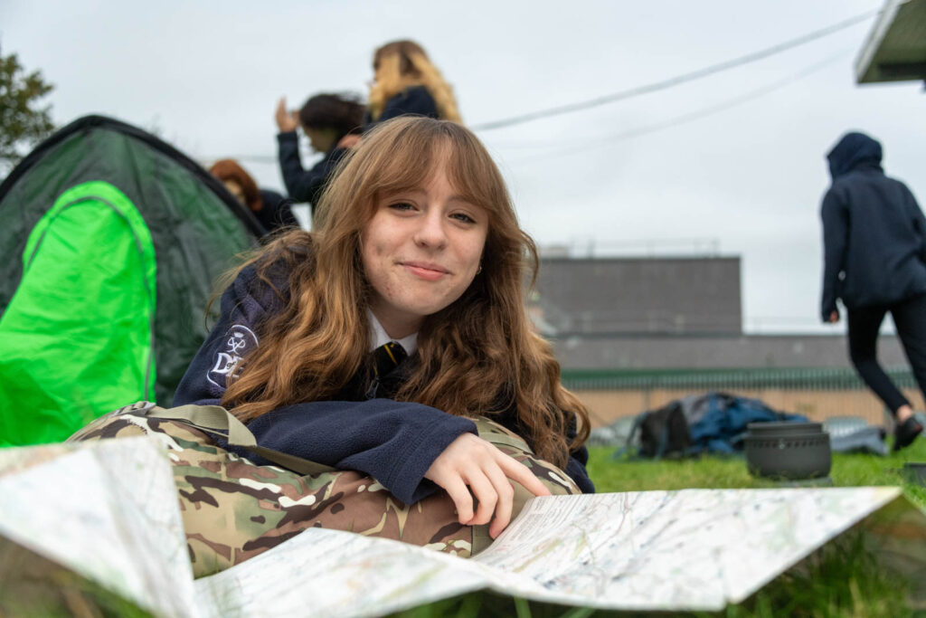 Pupil lying down on backpack and grass, looking at a paper map. Tent and pupils in background.