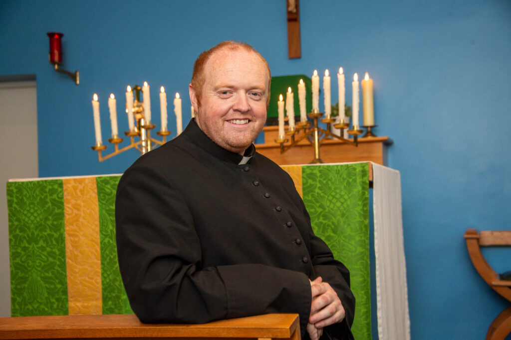 Priest sitting down and postering looking forward in front of altar. Lit candles and icons on altar and shelves.