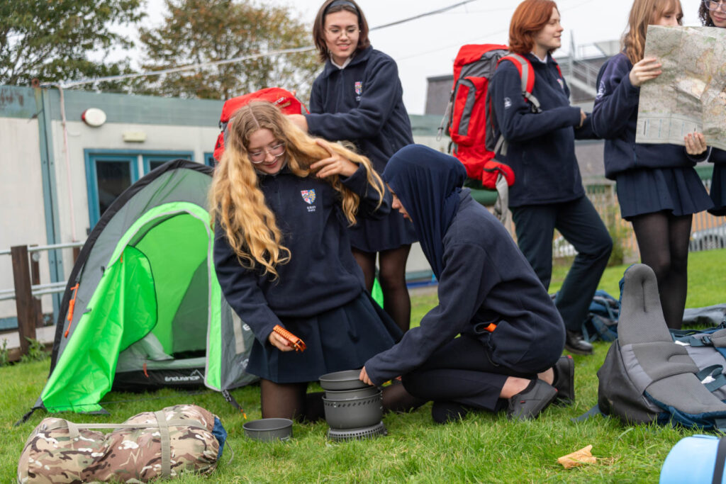 Pupils testing and setting up camp equipment and accessories, on school grass grounds. Maps, gas stoves, backpacks and tents. Some pupils looking up maps and others setting up a gas stove.