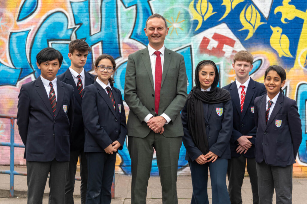 Staff member Mr Clinton, Headteacher with pupils, in front of school graffiti mural, of crest emblem. Pupils iv'er side of Mr Clinton.