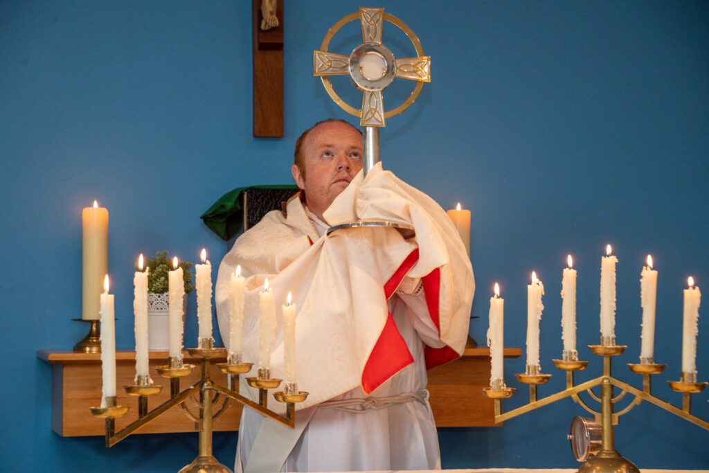 Chaplaincy priest performing Mass, holding up the cross. Surrounded by Catholic icons and lit candles. Cross with Jesus Christ on wall behind the priest.