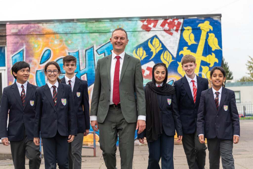 Staff member Mr Clinton, Headteacher, in front of school graffiti mural, of crest emblem. Walking forwards along side with pupils iv'er side of Mr Clinton.