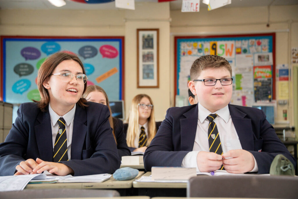 Class of pupils sitting down at desk's in classroom with open books looking forward to front of classroom.