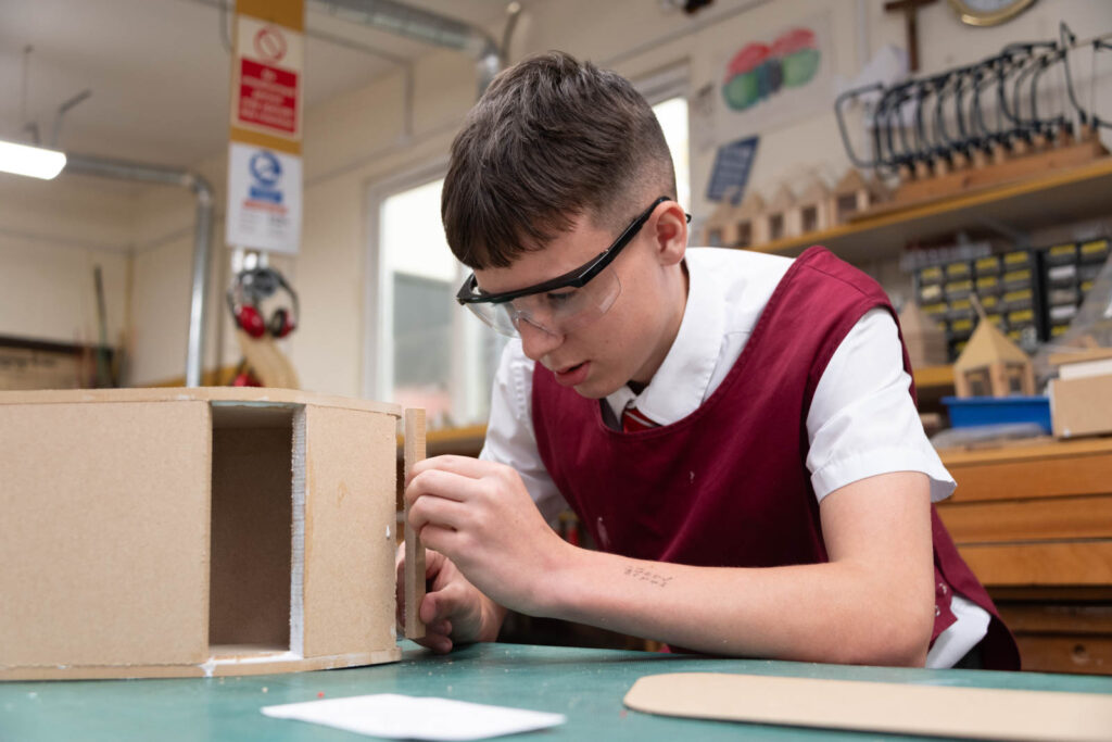 Pupil with safety equipment on in Dt class room workshop glueing a wooden project together.