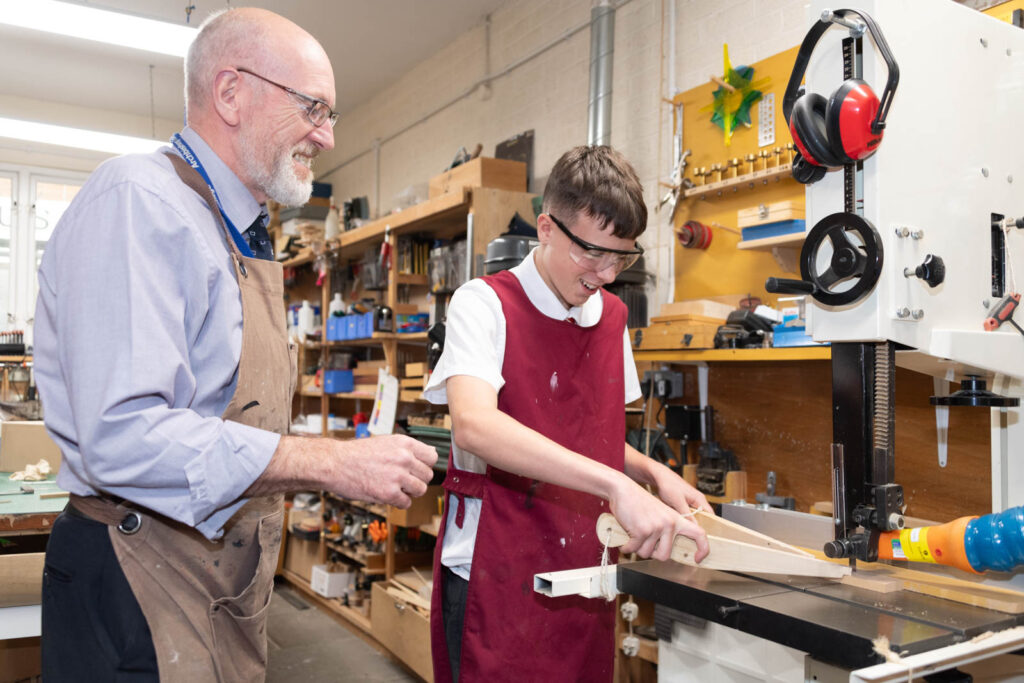 Pupil with safety equipment on using a bandsaw in DT class workshop to cut wood. While teacher helps supervise.