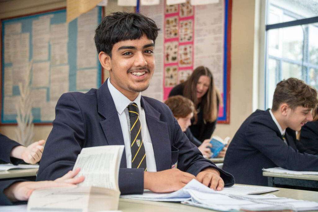 Class of pupils sitting down at desk's in classroom with open books in hands, reading. Teacher helping pupil's in background.