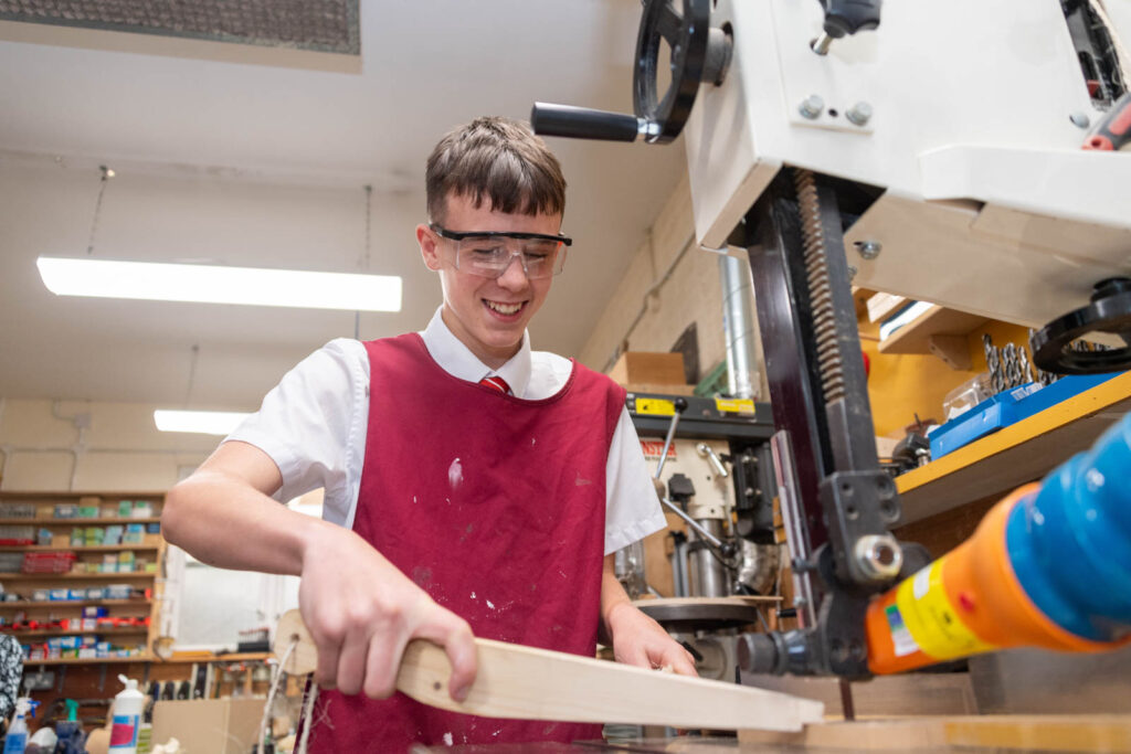 Pupil with safety equipment on using a bandsaw in DT class workshop to cut wood.