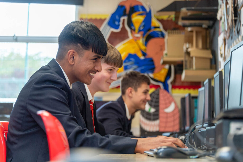 Pupils smiling at desks learning on computer. Art work on walls and models on shelves.