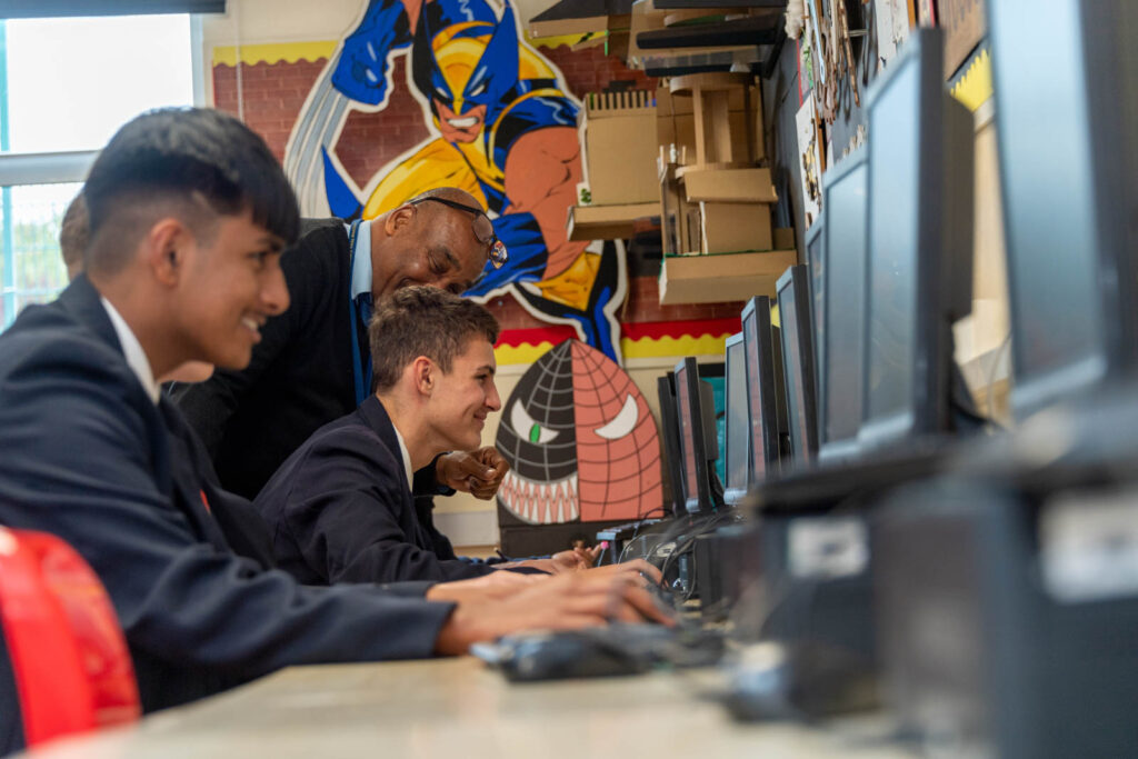 Pupils at desks learning on computers while teacher helps and is talking to a pupil. Art work on walls and models on shelves.