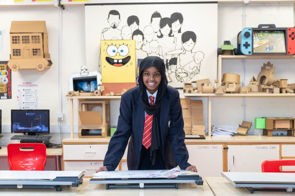 Pupil looking directly ahead leaning over work, in the DT classroom. in background on desk and shelves, student made models and art. Desktop computer also on table.