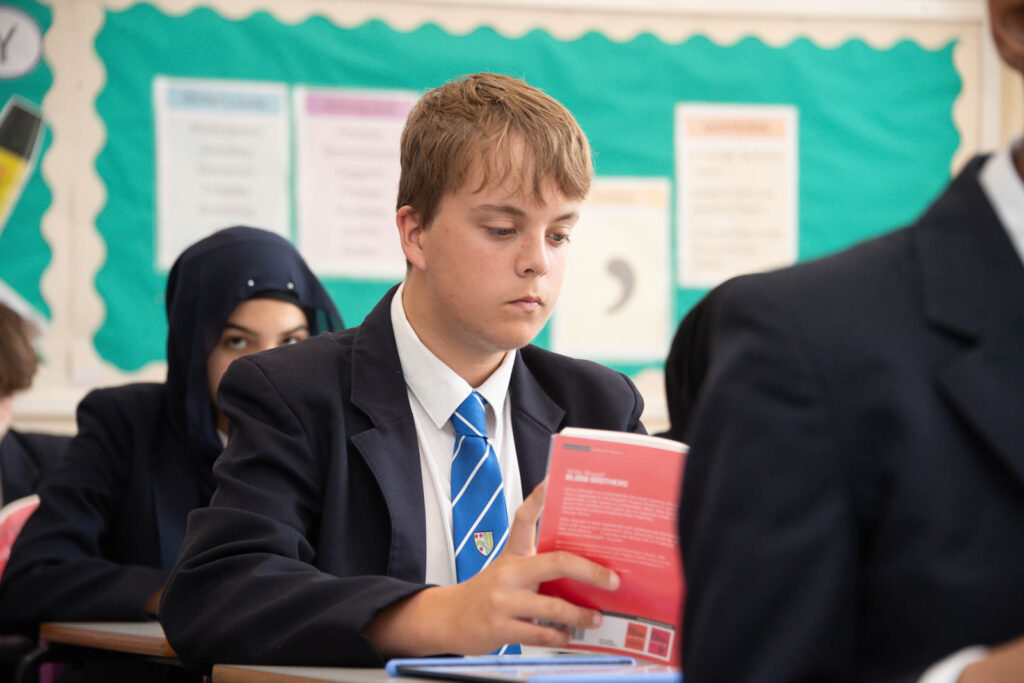 Full class of pupils sitting down at desk's in classroom with open books in hands, looking down reading the book.