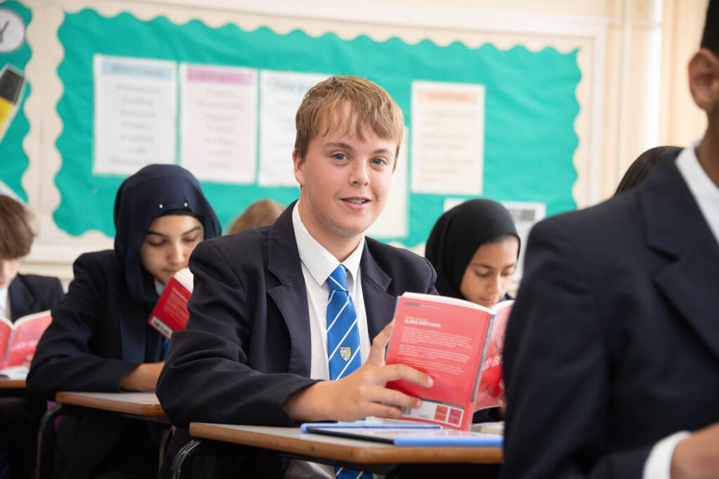 Full class of pupils sitting down at desk's in classroom with open books in hands, looking down reading the book. One pupil looking direct ahead.