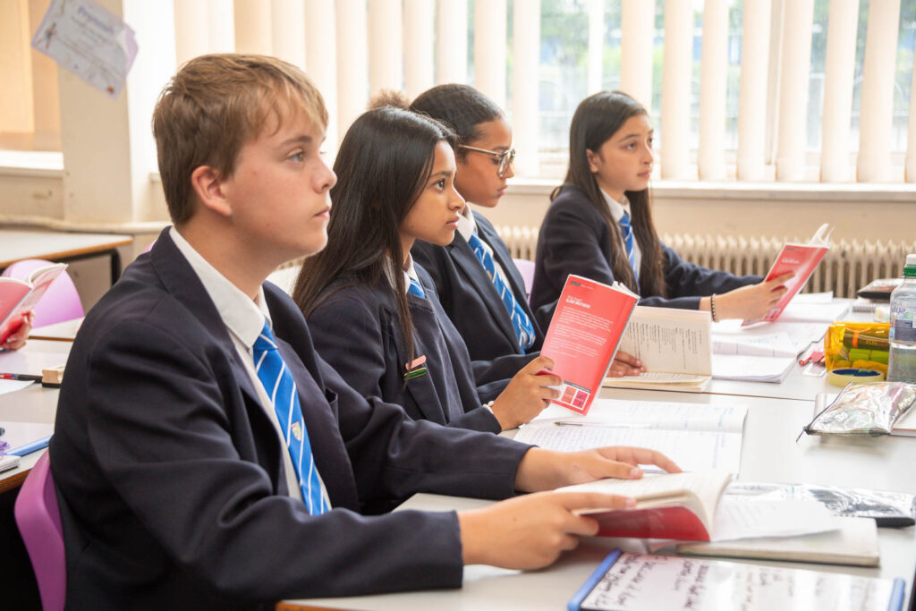 Pupils sitting down at desk's in classroom with open books in hands, looking towards front of classroom.