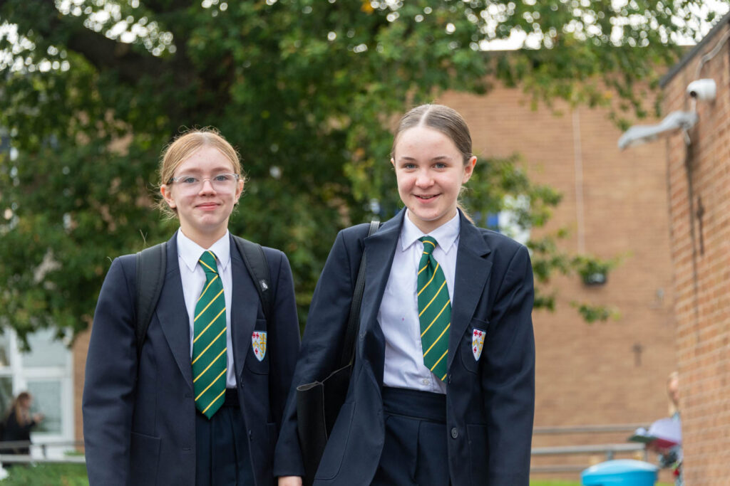 Two pupils outside on school grounds walking looking directly forward ahead smiling. School building's and tree in background.