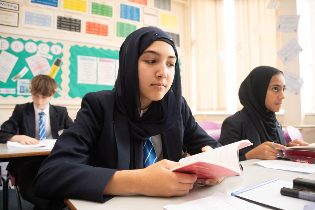 Two pupils with books in hands open , looking forward ahead at front of classroom. pupil in background on separate table/desk reading from book looking down. Back classroom wall decorated with informational posters and cut out paper decorations.