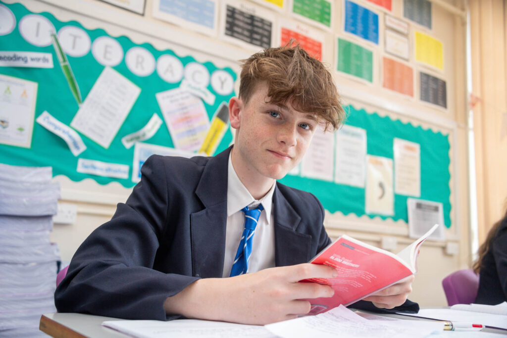 School pupil/student in classroom at desk holding open a red book looking direct. Classroom wall in background decorated with posters and some hand craft paper designs.