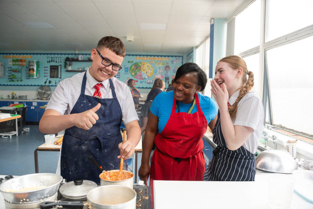 Pupils/Students learning to cook in lesson the with teacher in kitchen classroom. Everybody happy smiling with one pupil putting thumb up.