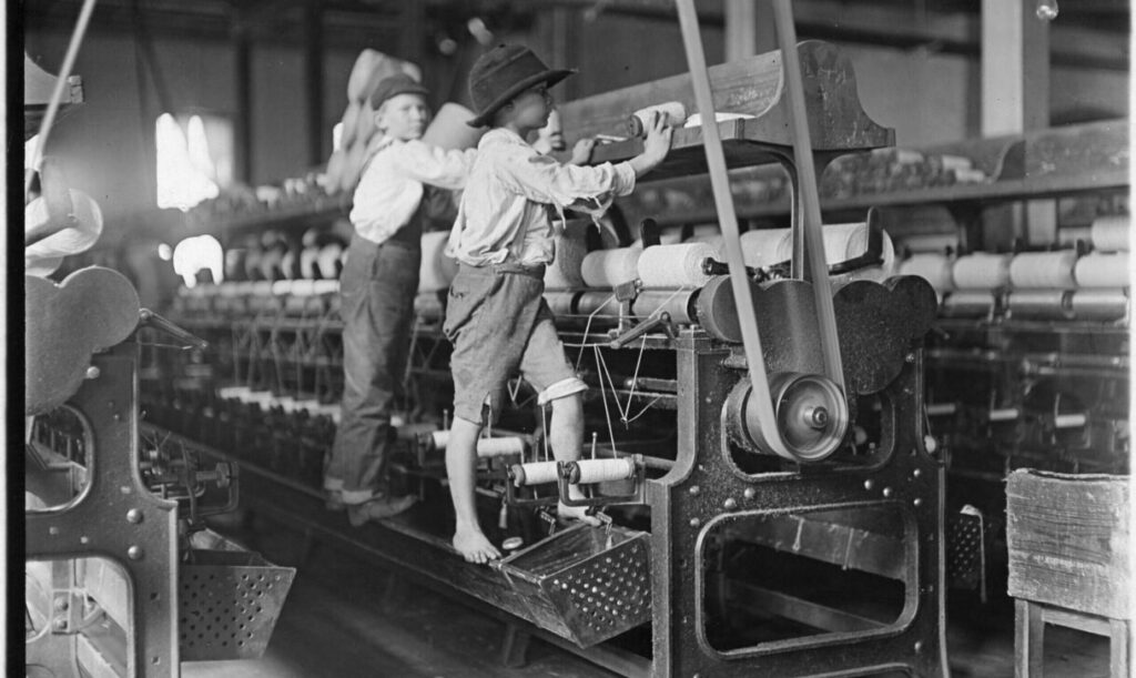 Many youngsters here. Some boys and girls were so small they had to climb up on to the spinning frame to mend broken threads and to put back the empty bobbins, January 1909. Photographer: Hine, Lewis.