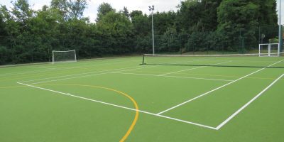 School's outdoor astro turf football pitch, tennis court, with flood light and trees in background.